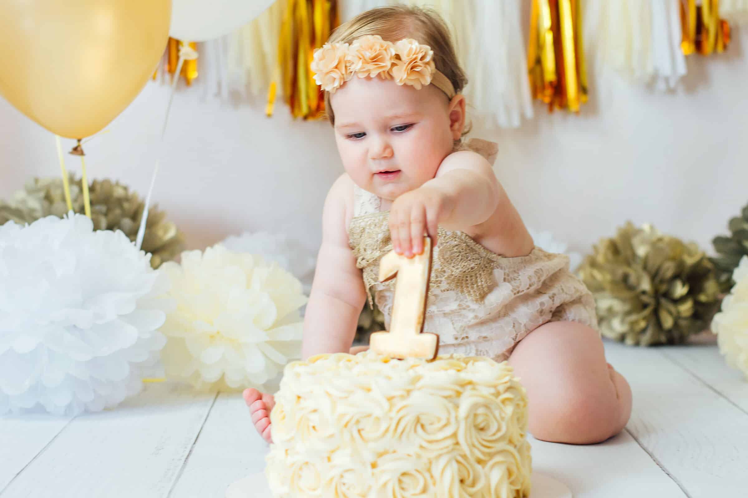 Baby in a flower headband reaching for the gold number 1 on a frosted smash cake