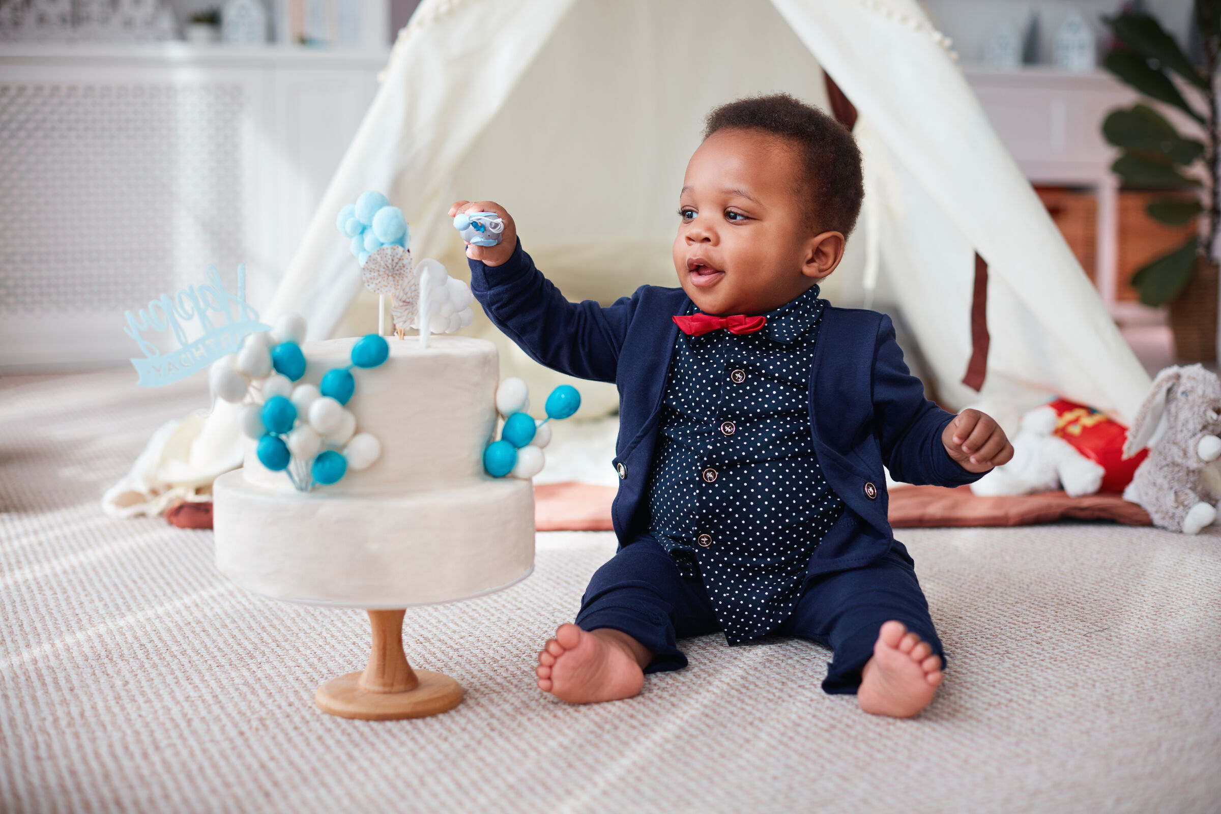 Toddler in a small suit reaches for his birthday cake at a Chuck&nbsp;E.&nbsp;Cheese party room with a play tent and stuffed toy.