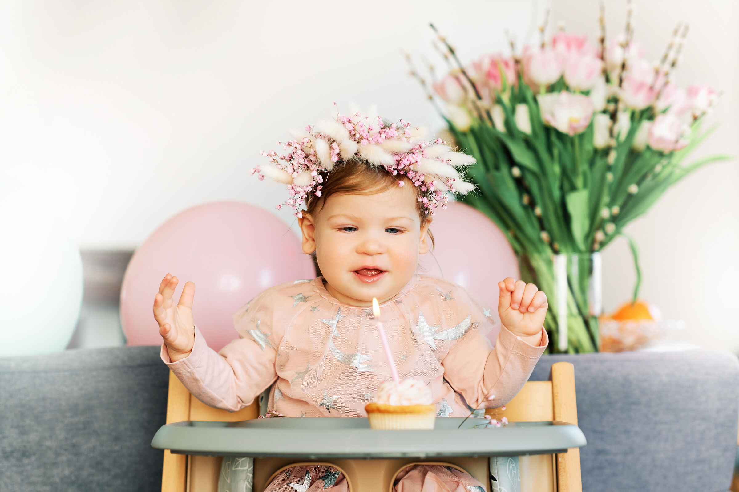 Toddler sits in a high chair next to a cupcake with a candle in a Chuck&nbsp;E.&nbsp;Cheese party room with pink balloons.