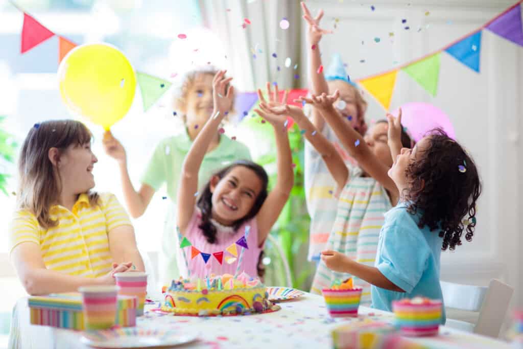 Kids ages 5–8 celebrate a birthday at Chuck E. Cheese party room table with cake, balloons, confetti, and pizza.