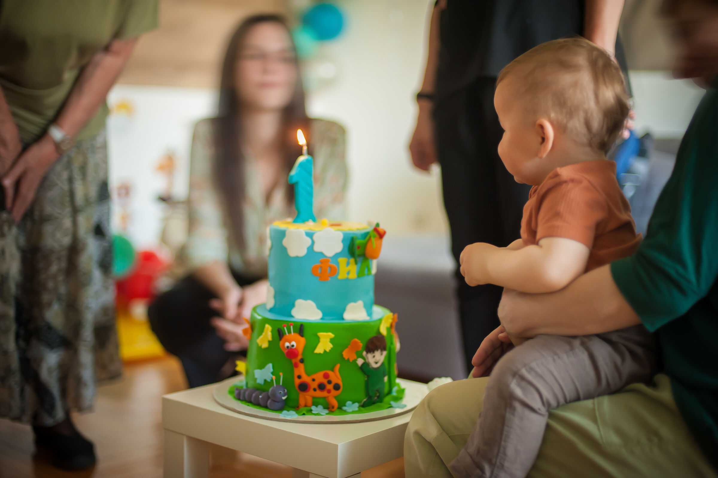 A parent holds a toddler at a Chuck&nbsp;E.&nbsp;Cheese party room with a single-candle cake decorated with clouds and animals.