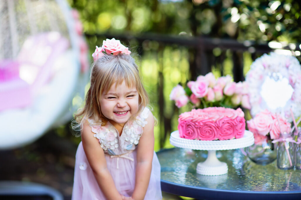 Birthday party at Chuck&nbsp;E.&nbsp;Cheese: girl (ages 4–6) in pink dress smiles by rose cake on glass table, party room with flowers.