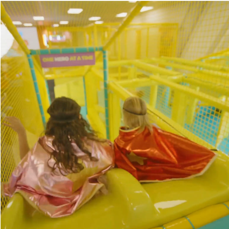 Two kids in capes and headbands sit atop a yellow indoor slide inside the Chuck&nbsp;E.&nbsp;Cheese Superhero Playground.
