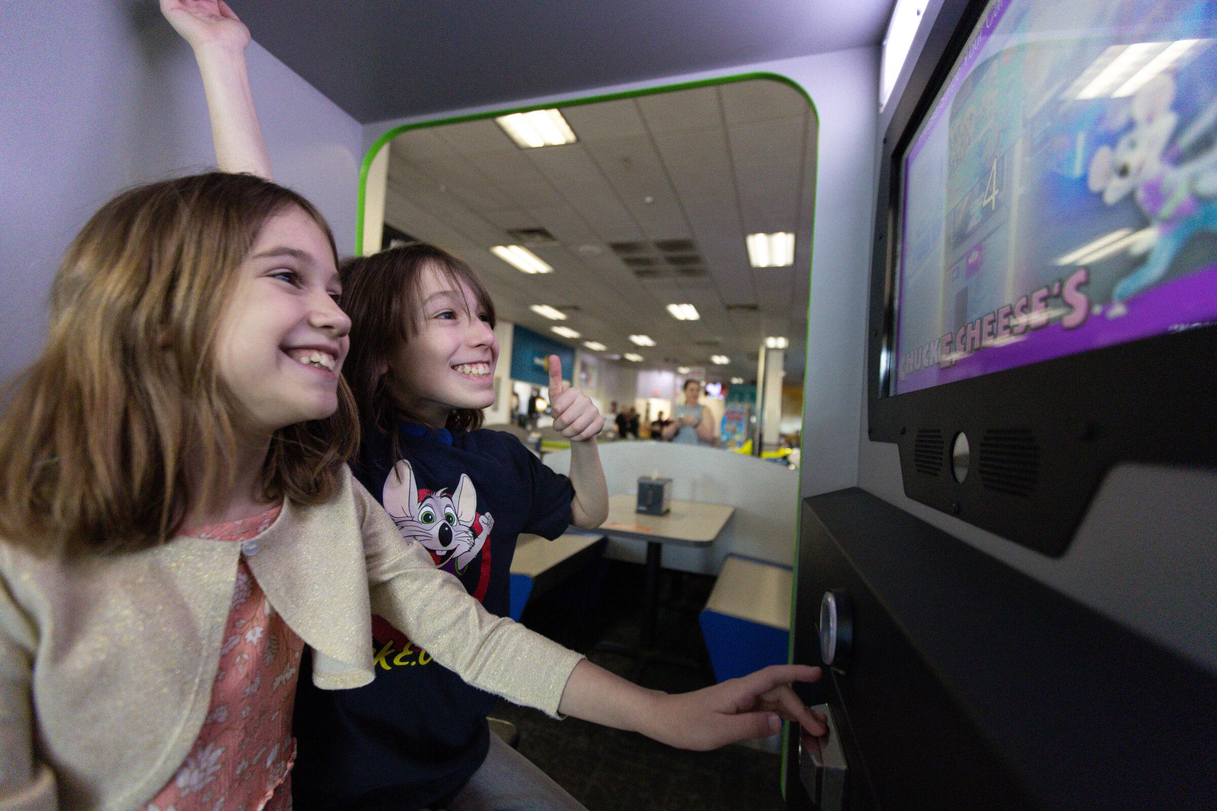 Two happy kids playing an arcade game at Chuck E. Cheese, one giving a thumbs up