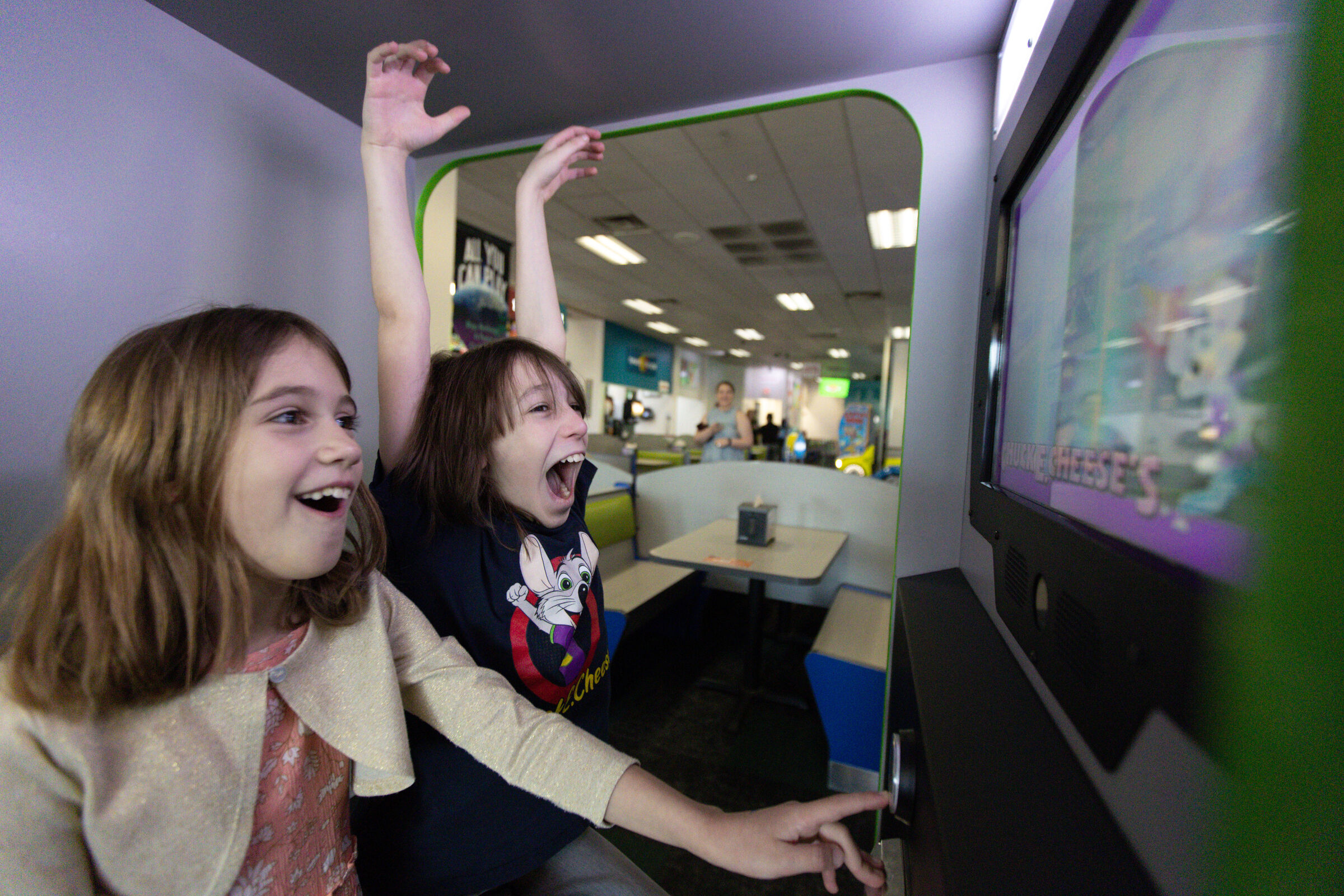 Two excited girls playing arcade games at Chuck E. Cheese, one cheering with arms raised
