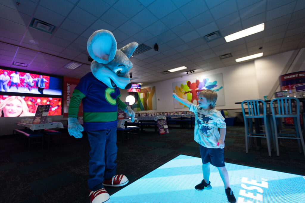 Chuck E. Cheese interacting with a young boy on a light-up dance floor.