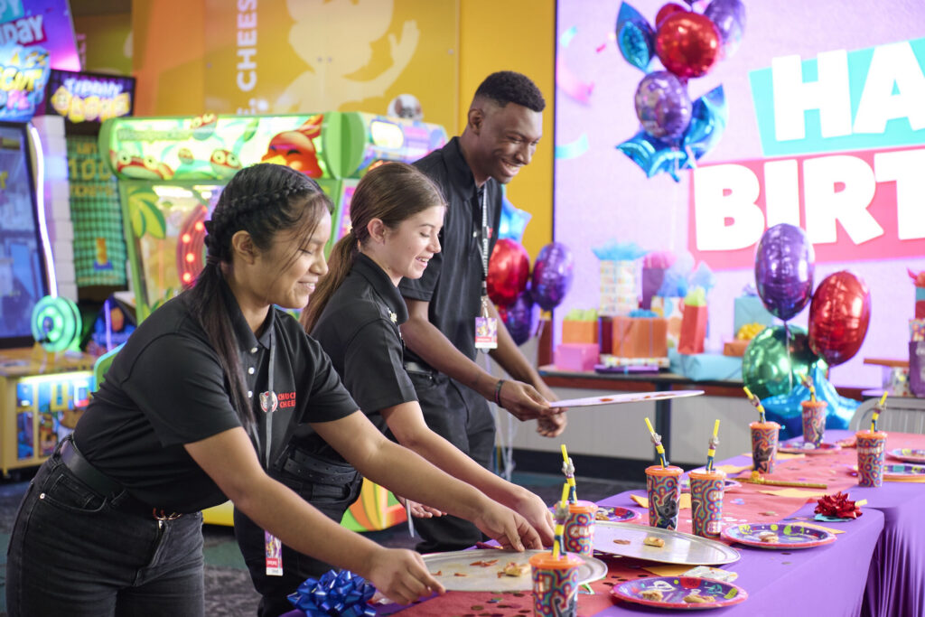 Chuck E. Cheese team members setting up a colorful birthday party table with pizza and decorations.