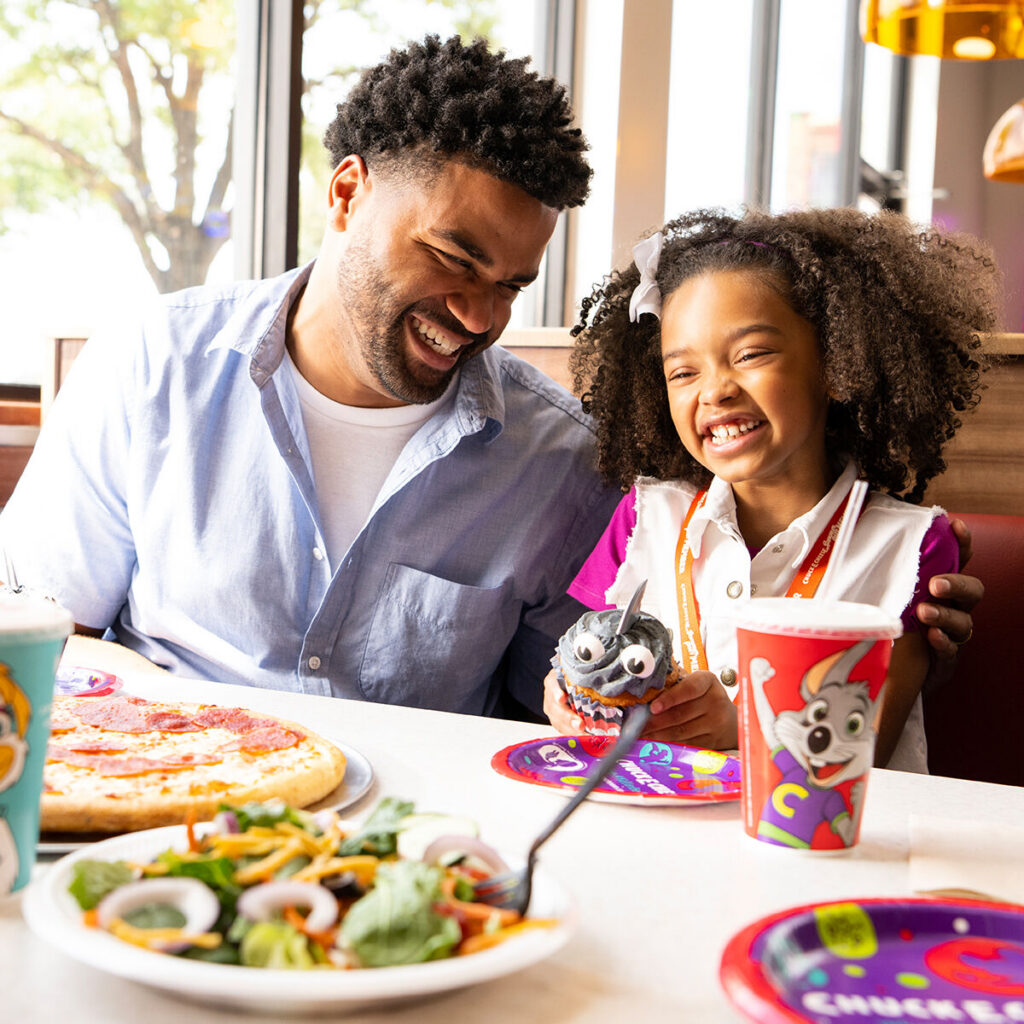 Father and daughter laughing joyfully while eating pizza and salad at a Chuck E. Cheese party table.