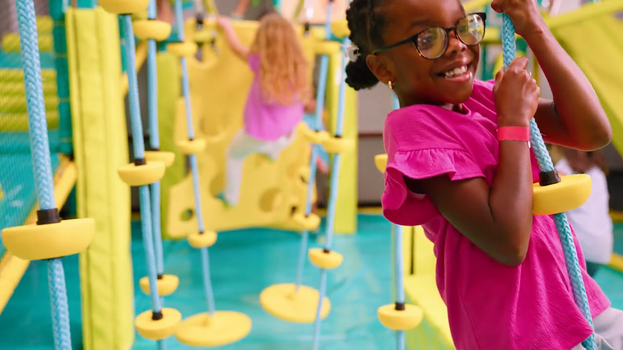 Girl with glasses smiling while climbing a rope ladder in the Chuck E. Cheese Adventure World play structure.