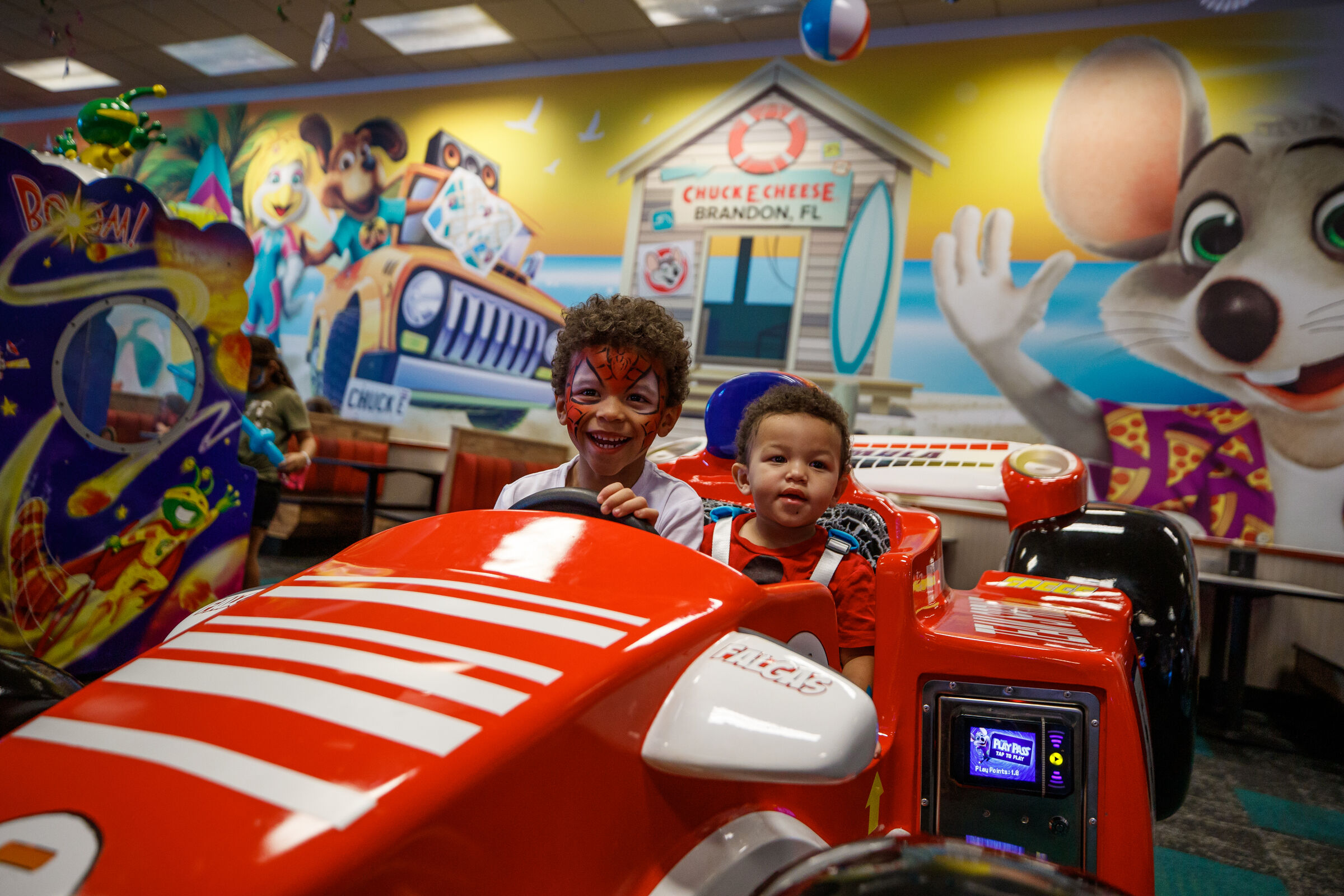 Two young boys smiling in a red race car arcade game at Chuck E. Cheese.