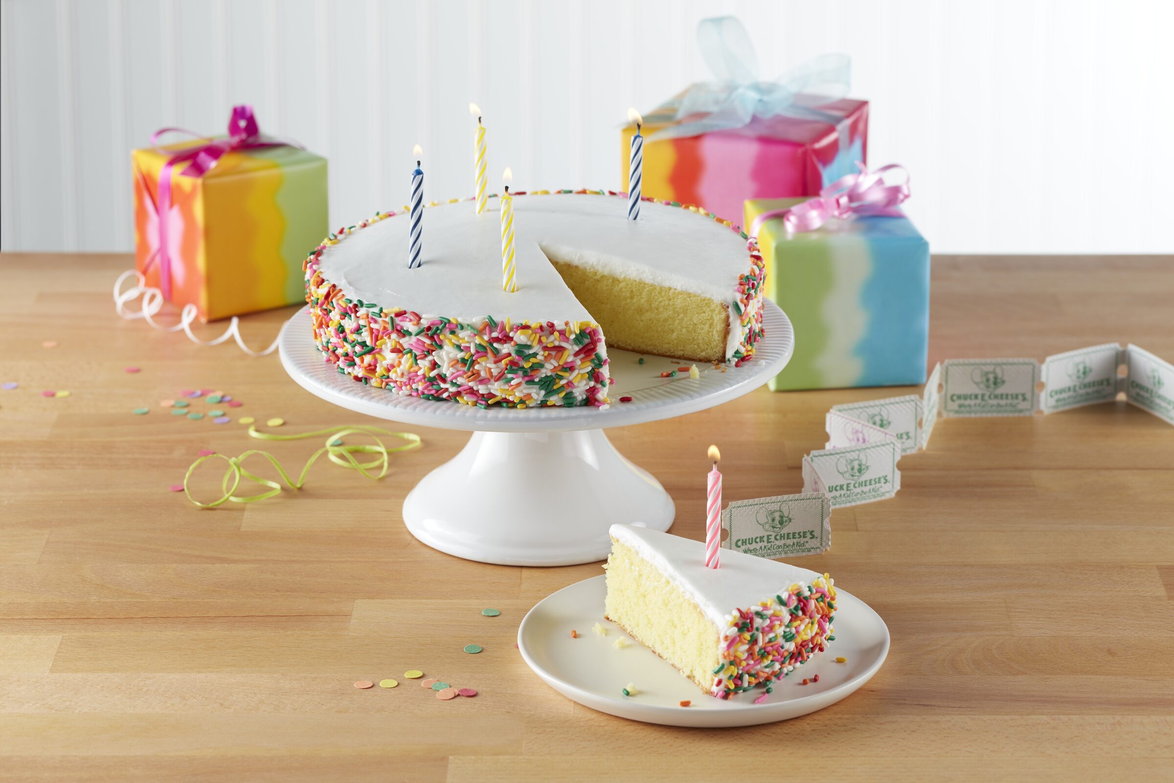 Birthday cake with four lit candles and presents in the Chuck E. Cheese party room.