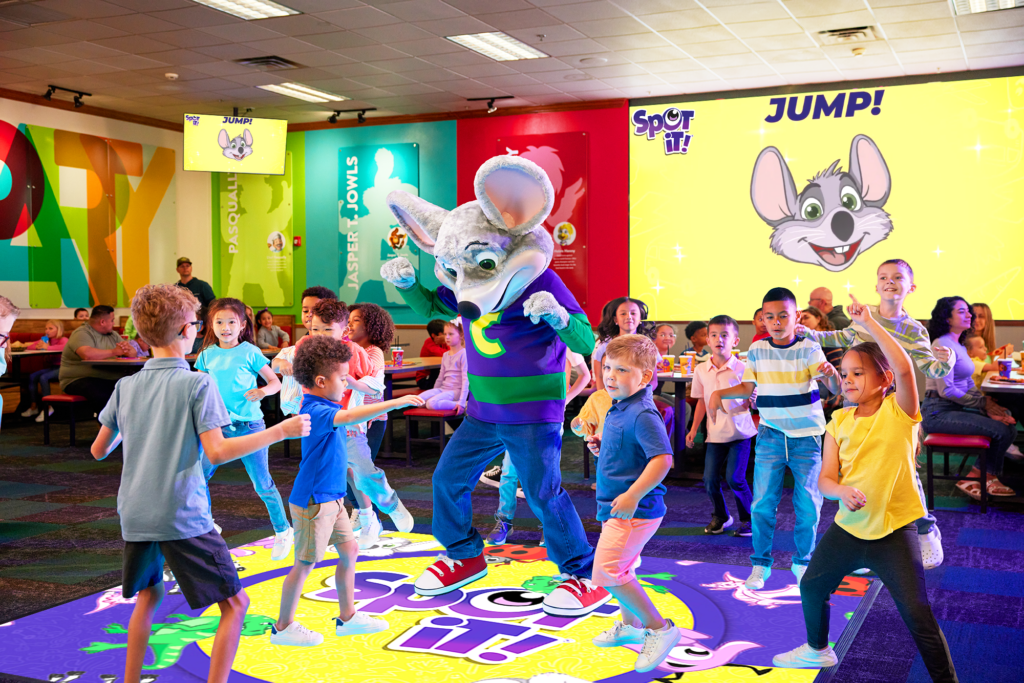 Chuck E. Cheese dancing with a group of kids on an interactive floor game at a Chuck E. Cheese location.
