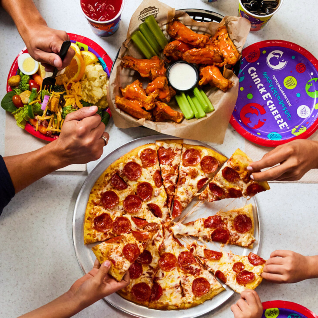 Family hands reaching for pizza, wings, and salad at a Chuck E. Cheese dining table.