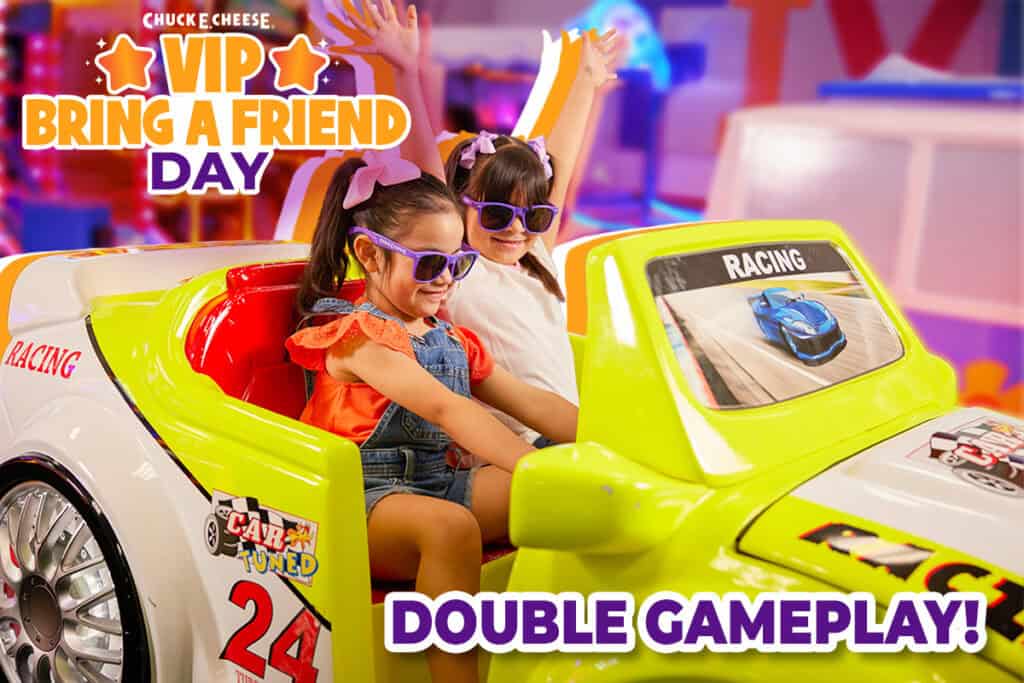 Two young girls wearing sunglasses and smiling while playing a racing arcade game at Chuck E. Cheese.