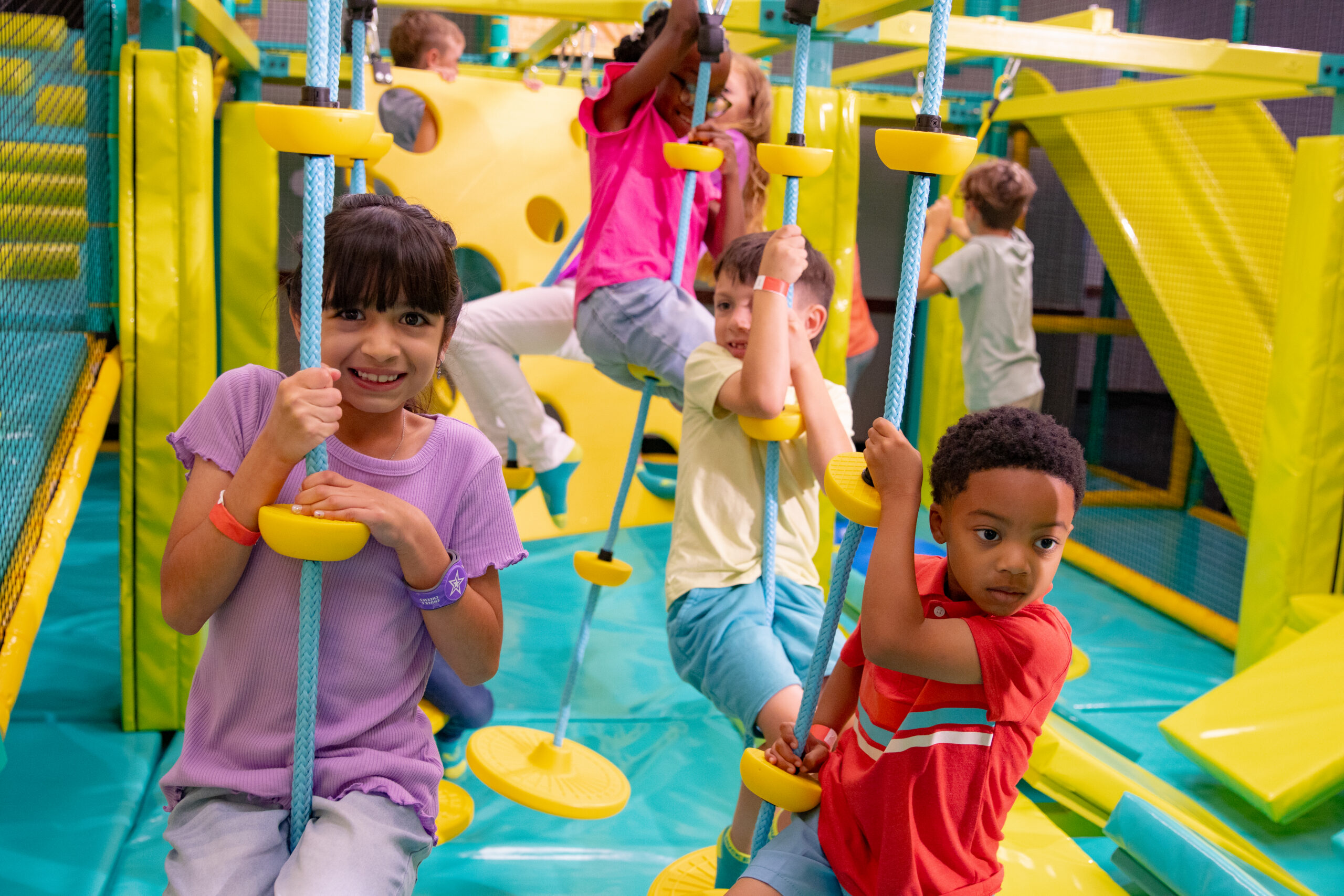 A toddler girl and older kids of different ages climb rope swings together in the Chuck E. Cheese indoor playground.