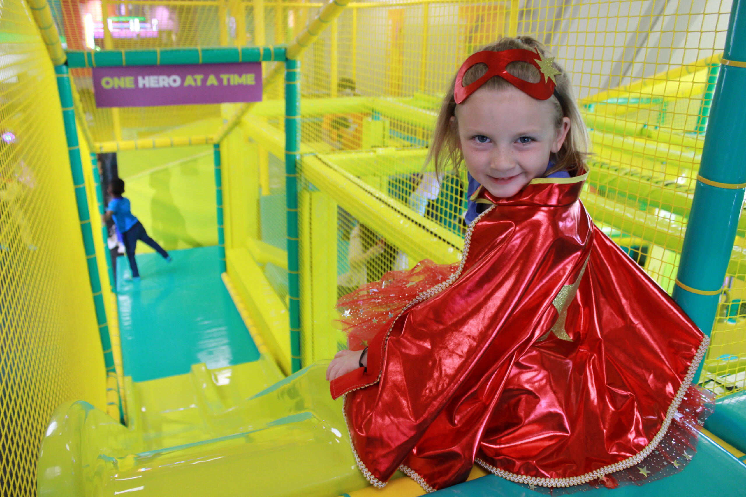 Girl in a superhero cape and mask smiling from inside the Adventure World play structure.