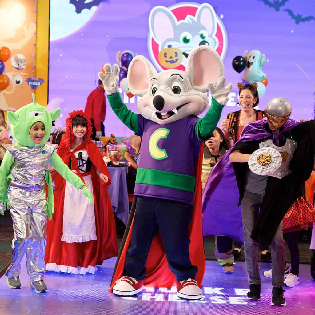 Chuck E. Cheese dancing with kids in Halloween costumes on a light-up stage at a Chuck E. Cheese Halloween party.
